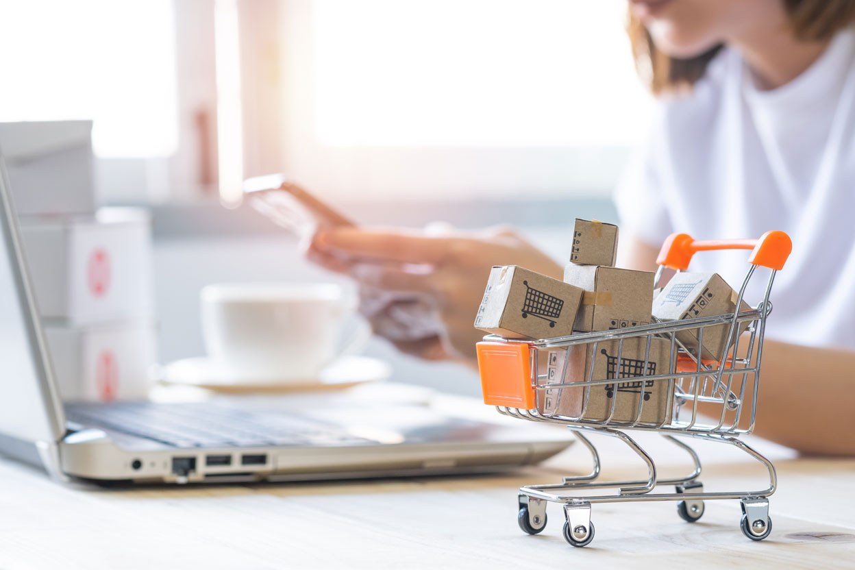 Blurry in the background, a woman holds her phone next to her computer. In front of her in focus is a shopping cart full of cardboard boxes with shopping carts on them