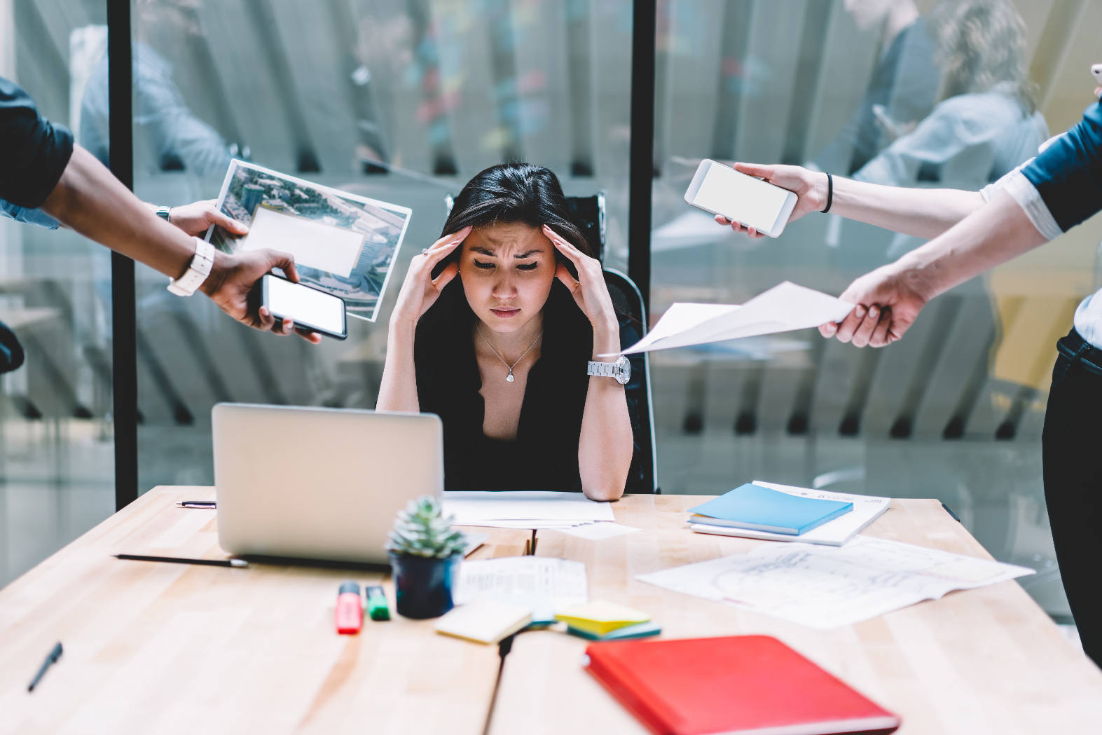 Une femme assise à un bureau, la tête entre les mains, est entourée de bras qui lui tendent des papiers