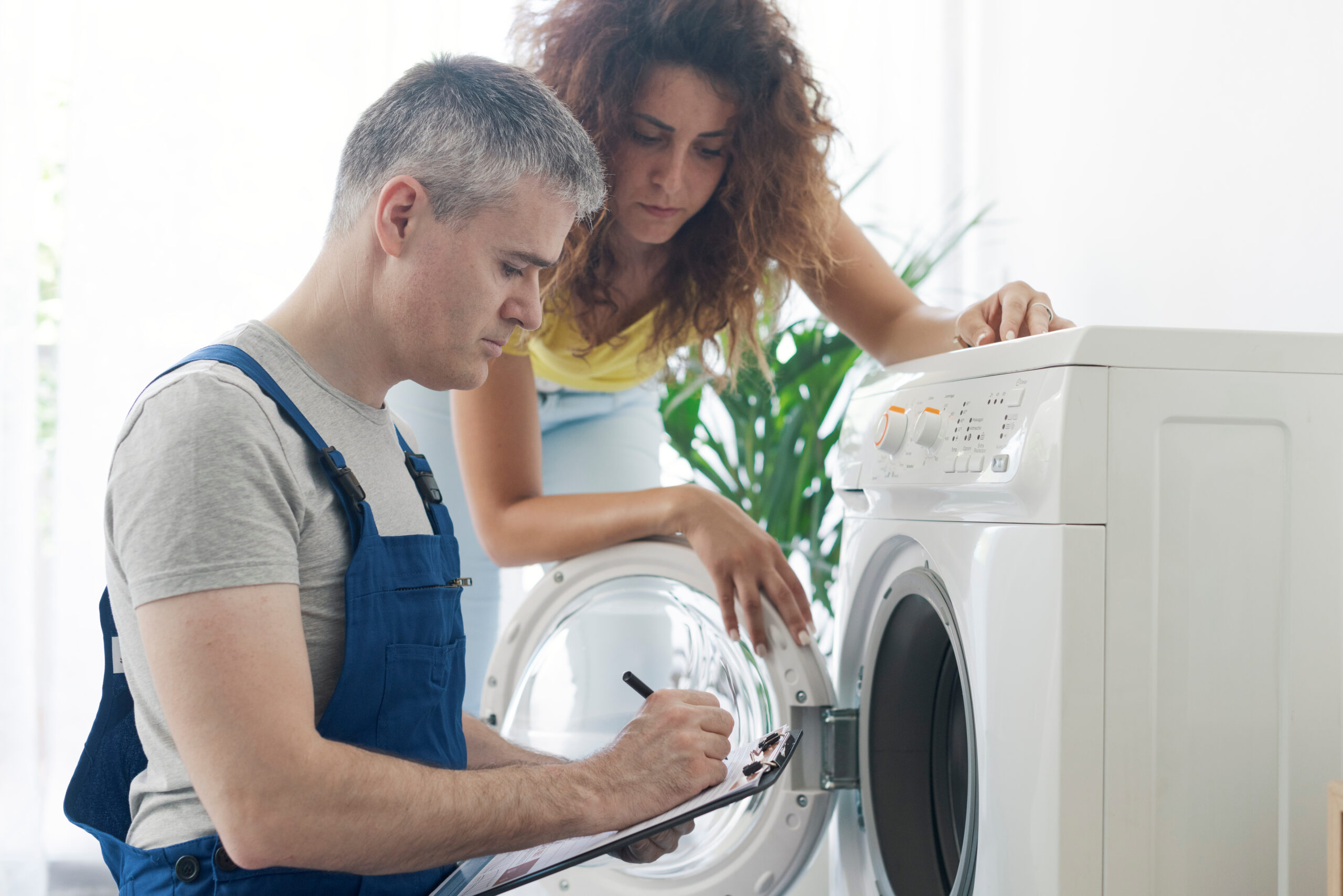 Technician performing a check-up on a washing machine, he is writing on a clipboard, repair service concept