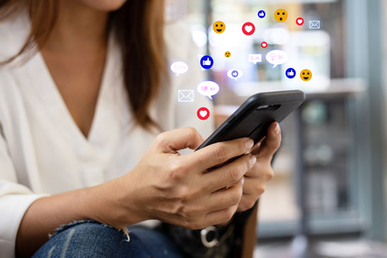 Une personne tient un téléphone dans les mains, au-dessus duquel flottent des emojis et des émoticones