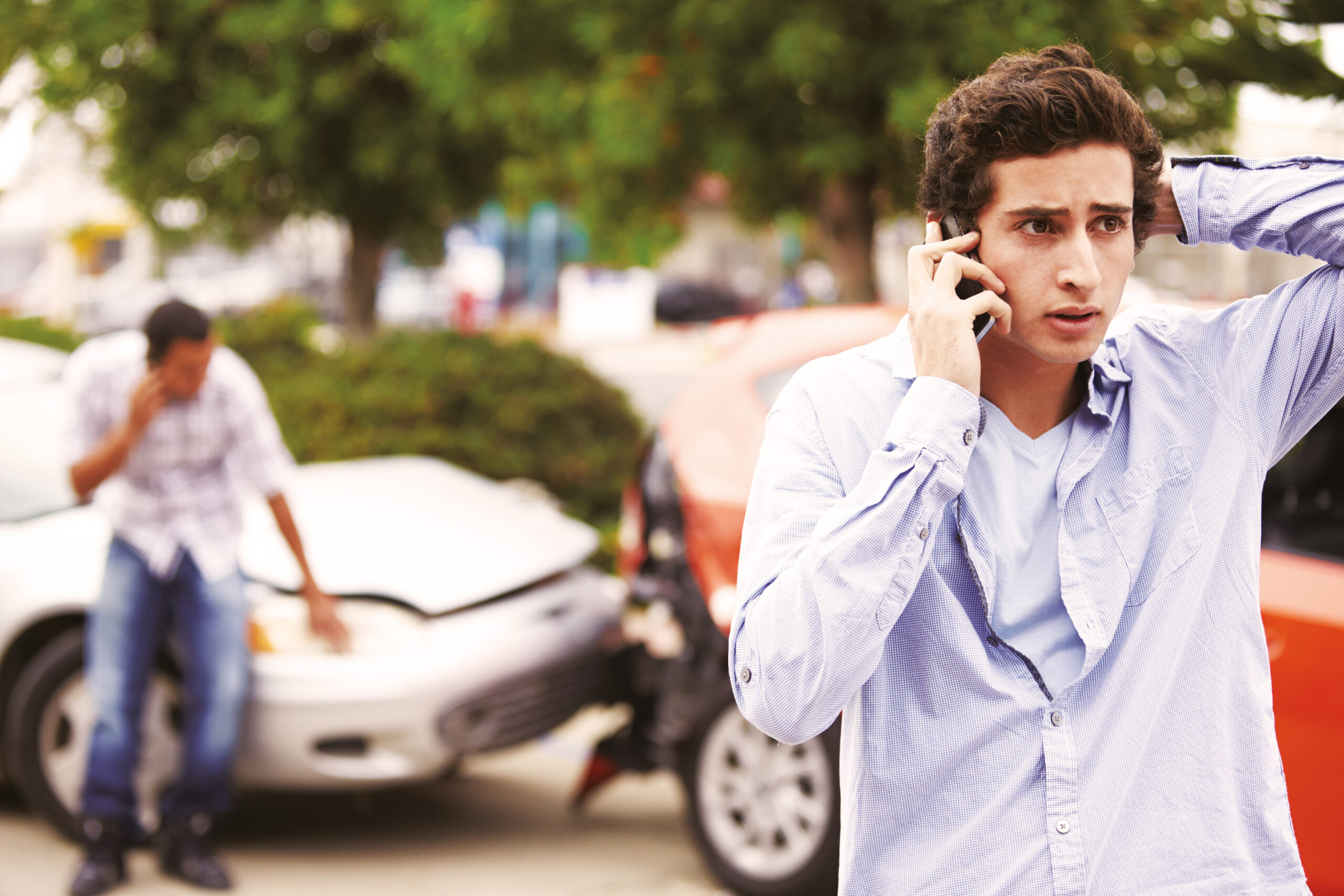 A man on the phone while another man in the background checks the two accidented cars