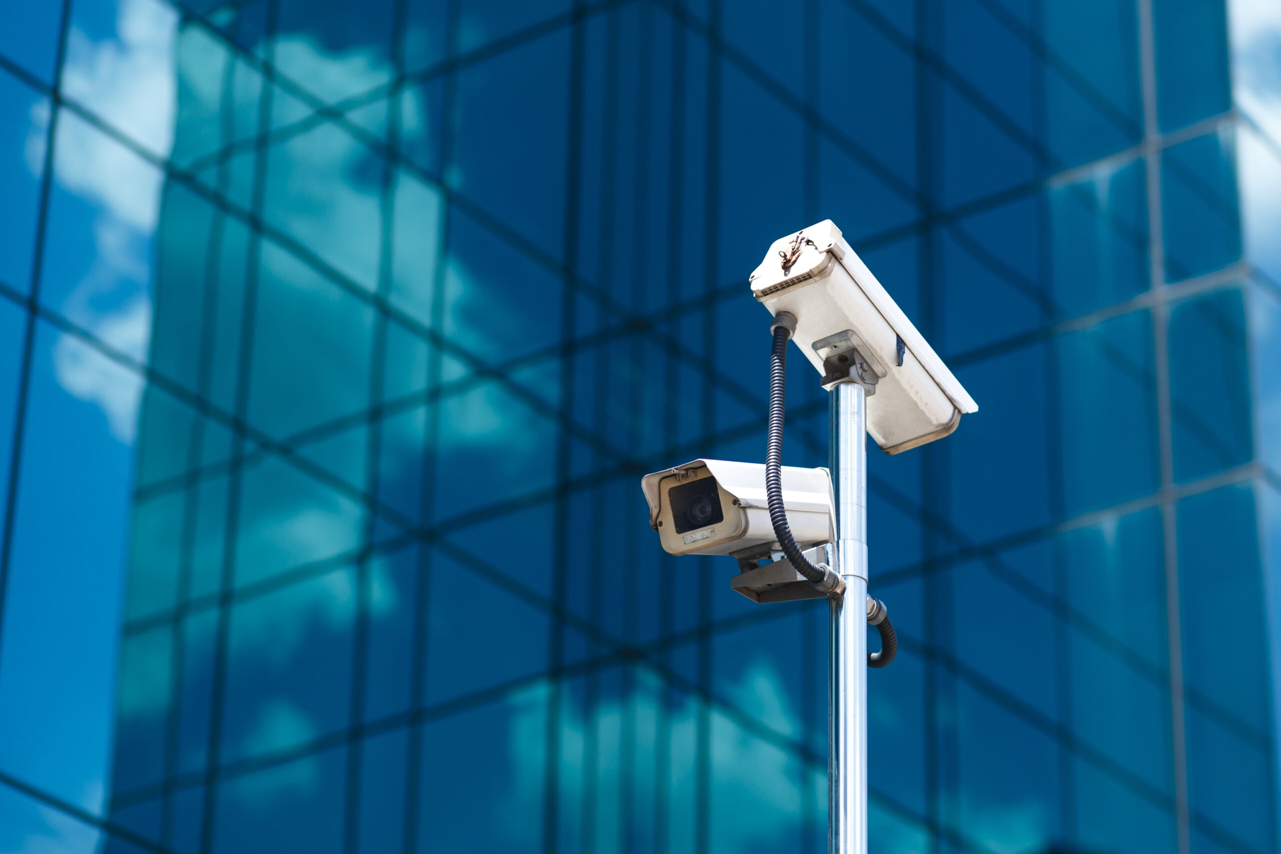 Un poteau surmonté de deux caméras de vidéosurveillance blanches, avec en arrière-plan un grand immeuble de bureaux aux façades vitrées.