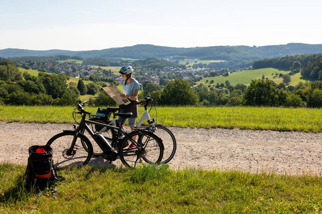 Une femme derrière deux vélos consulte une carte pour chercher son chemin. Elle est sur un chemin de terre surplombant une petite ville nichée au creux d'une vallée verdoyante.