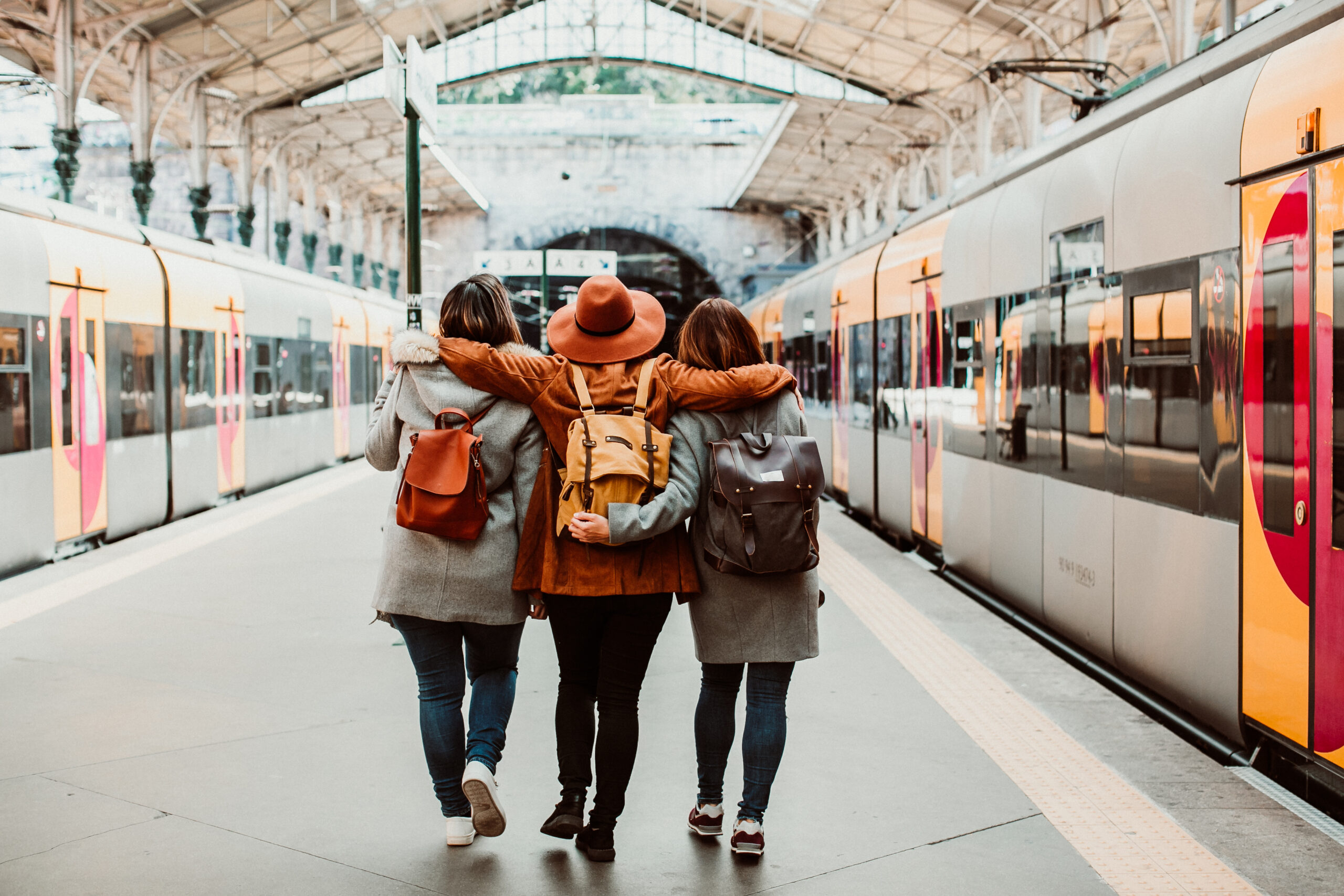 Trois femme avec des sacs à dos marchent le long d'un quai avec un train de chaque côté du quai. Elles sont proches et celle du milieu passe ses bras autour des deux autres.