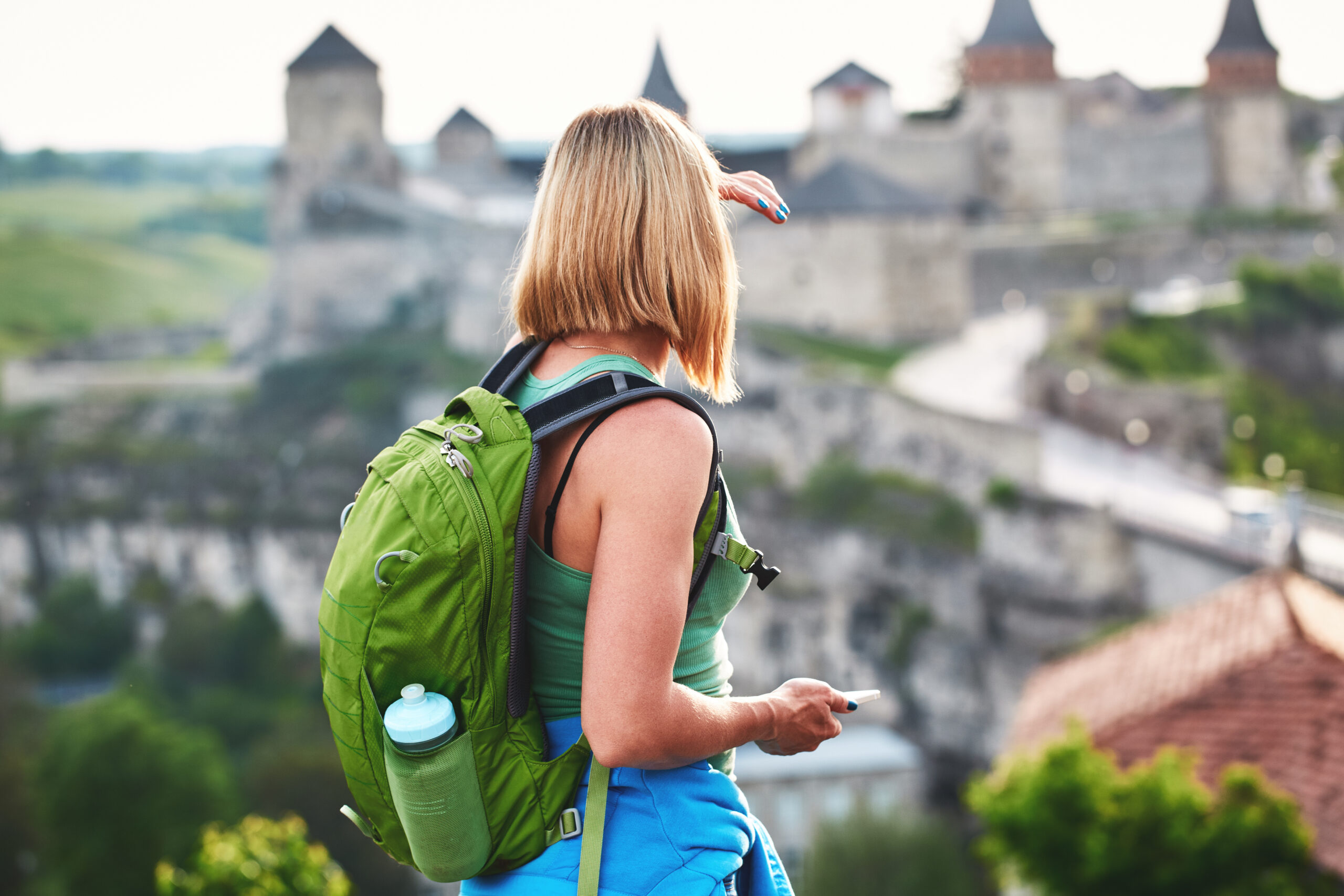 A tourist carrying a rucksack and a water bottle is gazing at a castle in the distance.