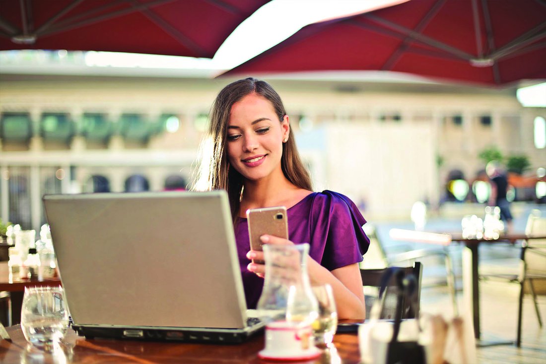 A woman looks at the screen of her smartphone, her laptop open in front of her.