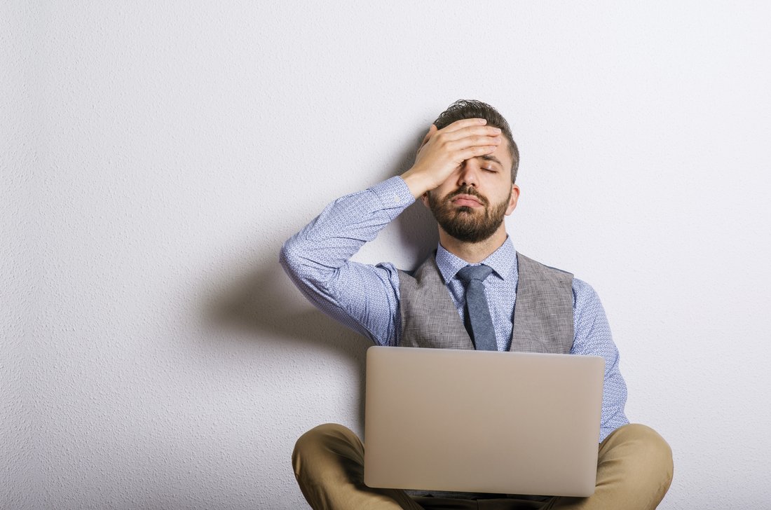 A man sitting with his laptop in his lap is holding his head with his hand.