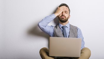 Un homme assis avec son ordinateur portable sur les genoux tient sa tête avec sa main.