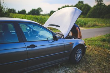 A man is repairing a car, with the hood open, on the side of a country road.