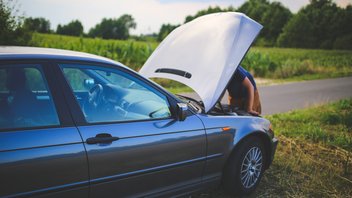 Un homme est en train de réparer une voiture, le capot ouvert, sur le bord d'une route de campagne.