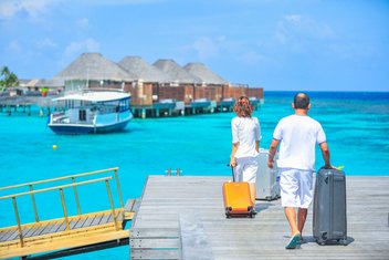  A couple walking suitcases by hand on a footbridge over turquoise water. Wooden and straw bungalows can be seen in the background.