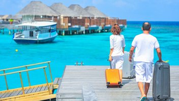  A couple walking suitcases by hand on a footbridge over turquoise water. Wooden and straw bungalows can be seen in the background.