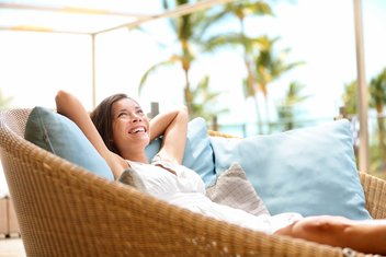 Une femme souriante est étendue au soleil, sur un grand fauteuil en osier rempli de coussins. On peut voir des palmiers en arrière-plan.