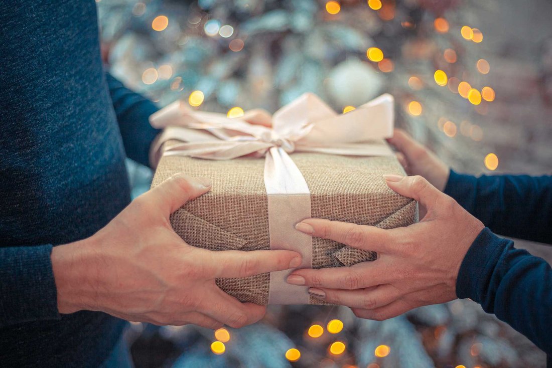 Un homme donne un cadeau emballé à une femme, avec un sapin de Noël illuminé en arrière plan.