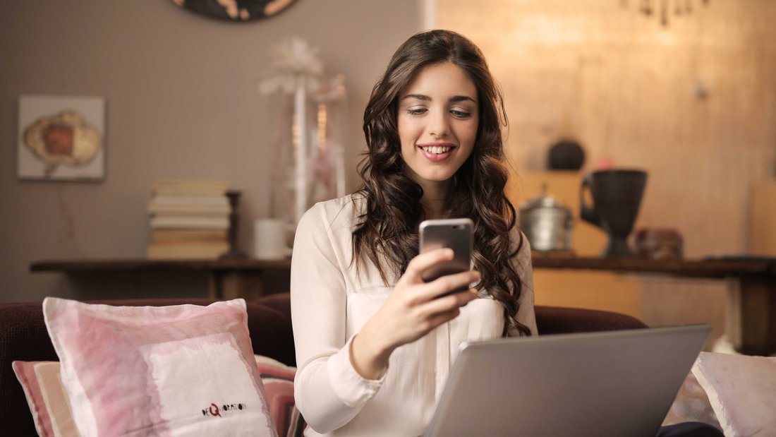 Une femme souriante assise sur un canapé tient son téléphone portable dans les mains et son ordinateur sur les genoux.