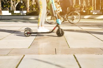 In the foreground, one person is riding a scooter, and another person in the background is riding a bicycle.