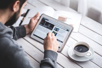 Un homme navigue sur sa tablette, une tasse de café à côté de lui.